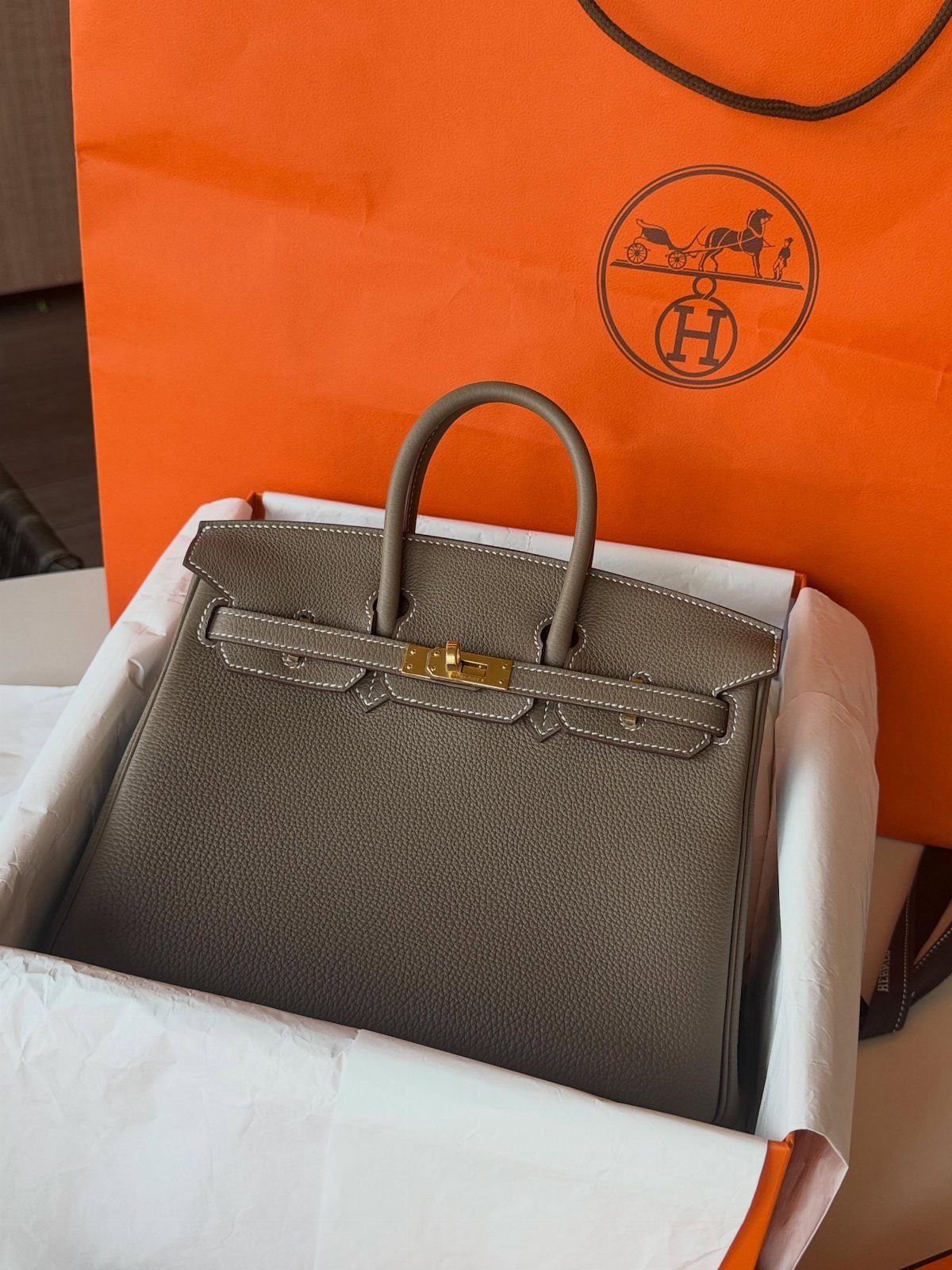 Grey leather handbag with gold hardware inside a white box and orange bag.