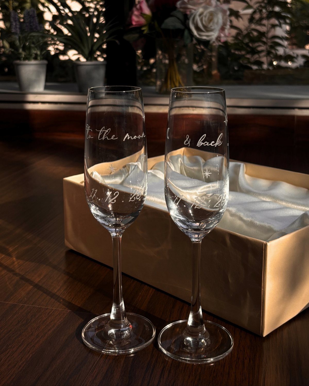 Two engraved champagne flutes standing before a gold gift box on wooden table