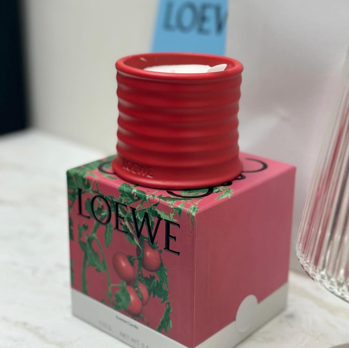 Red ribbed Loewe candle resting on a pink box with tomato illustrations.