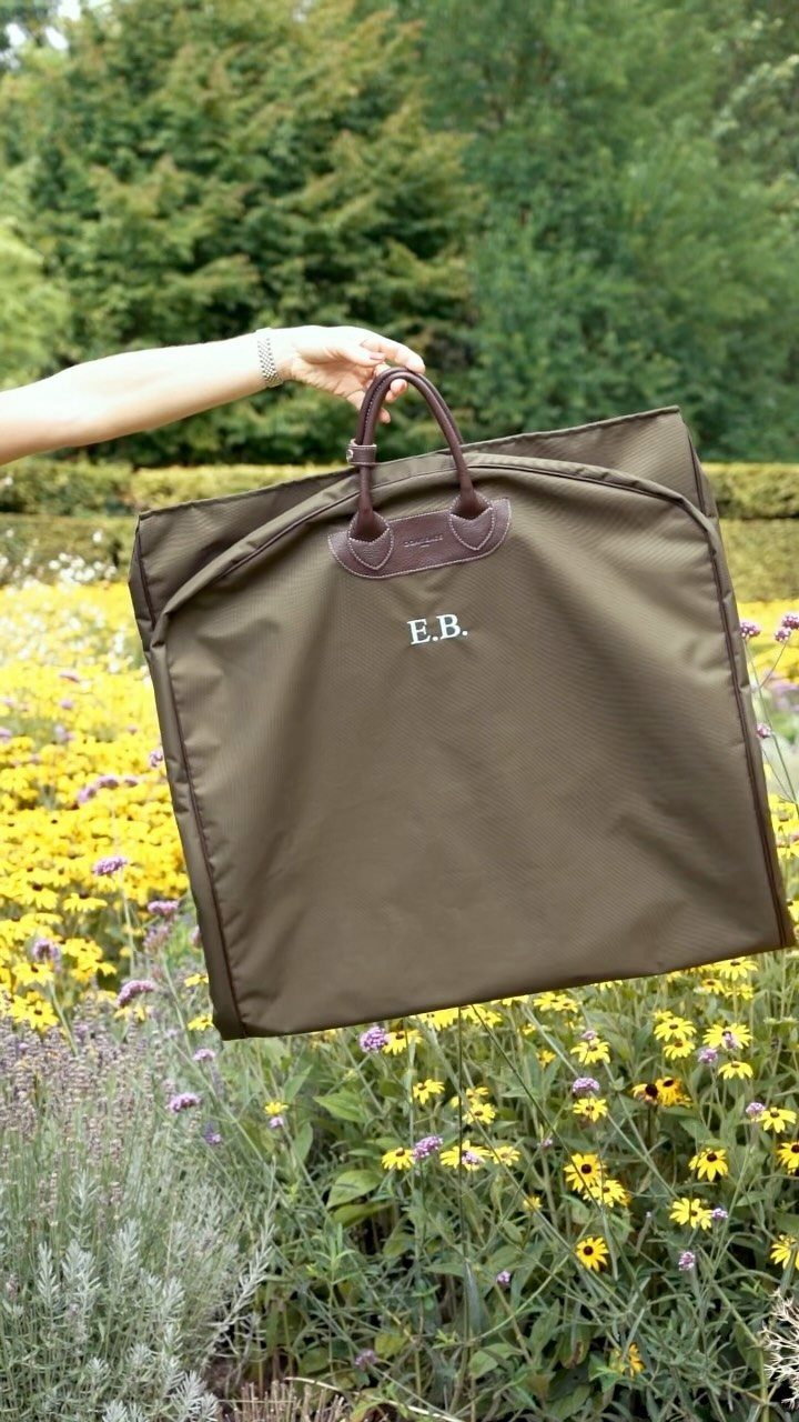 Brown monogrammed garment bag held over a field of yellow and purple flowers.