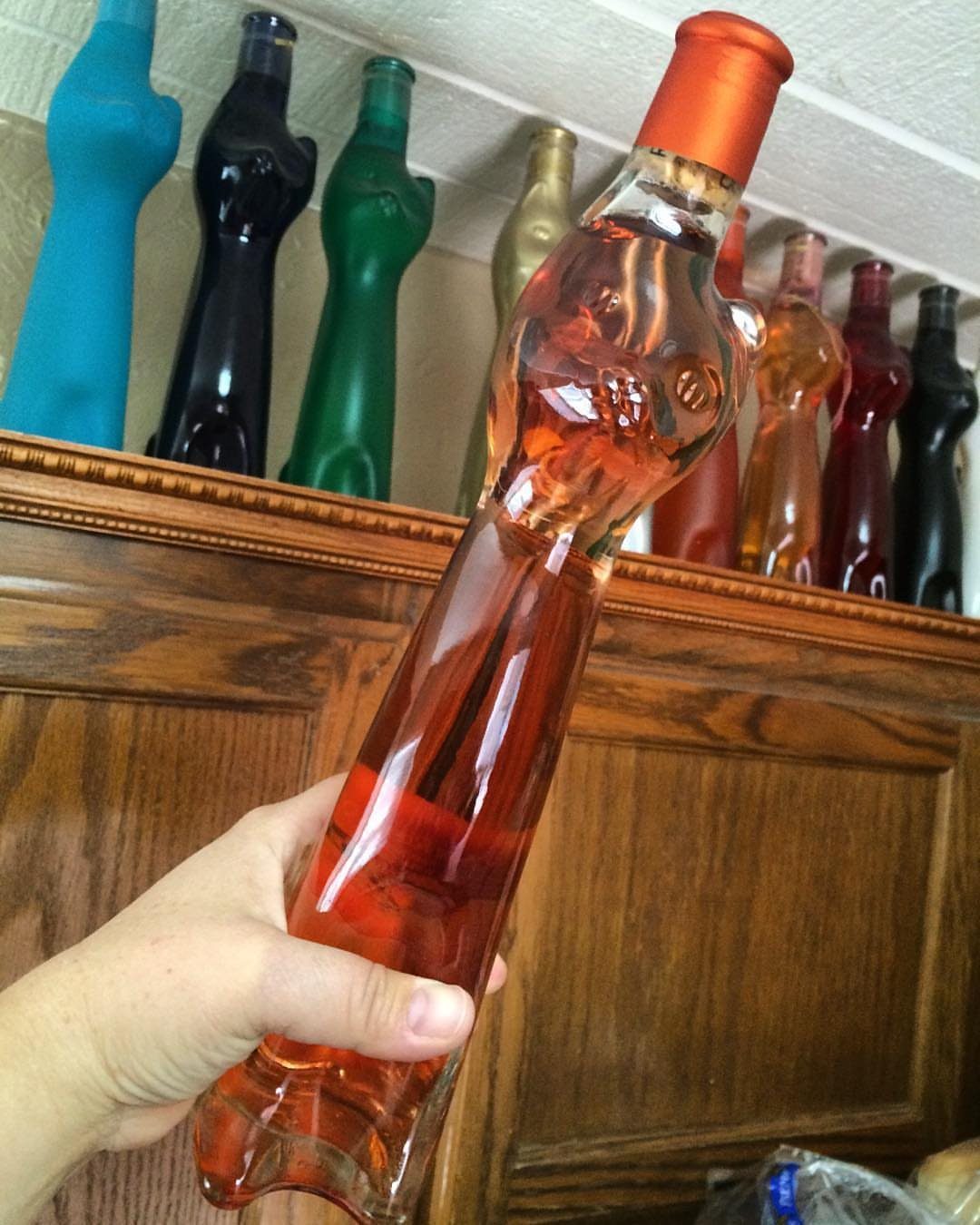 Human-shaped glass bottle filled with reddish-orange liquid held against a wooden shelf.