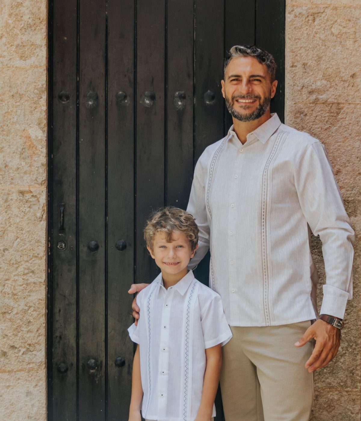 Man and boy wearing white guayabera shirts standing before a dark wooden door.