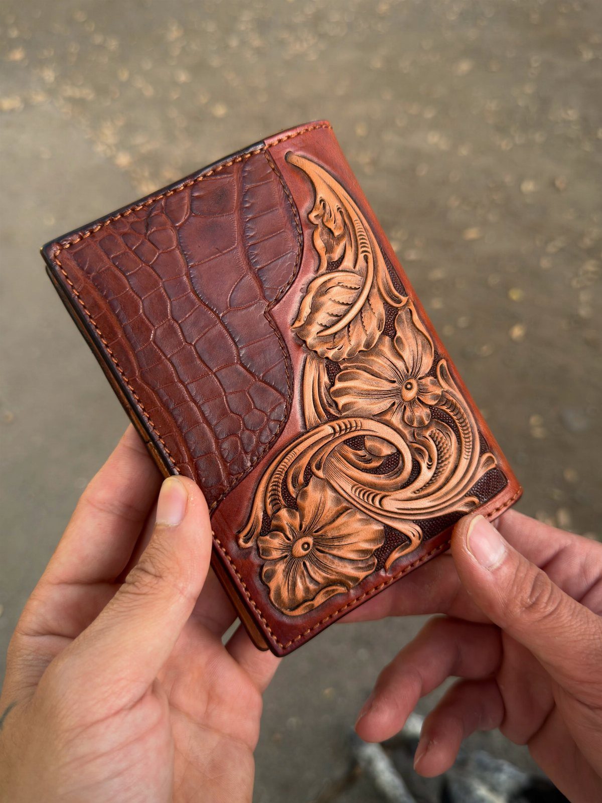 Handcrafted brown leather wallet with floral carvings and crocodile texture held by hands.