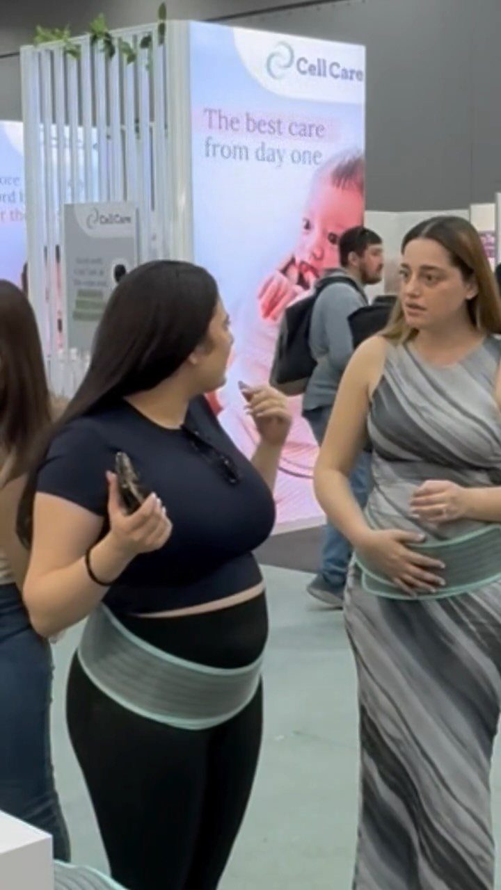 Two women wearing waist support belts while standing at a trade show booth.