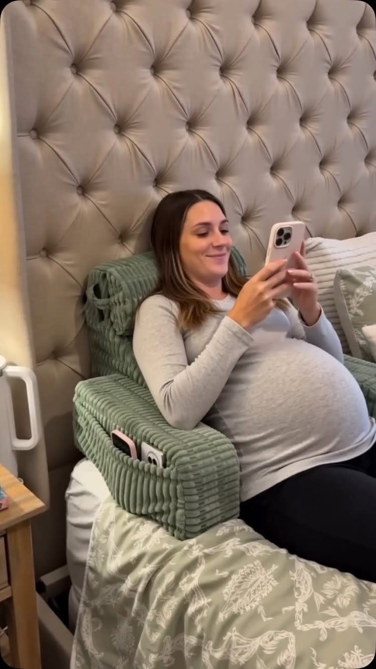 Pregnant woman sitting in bed supported by a large sage green pregnancy pillow.