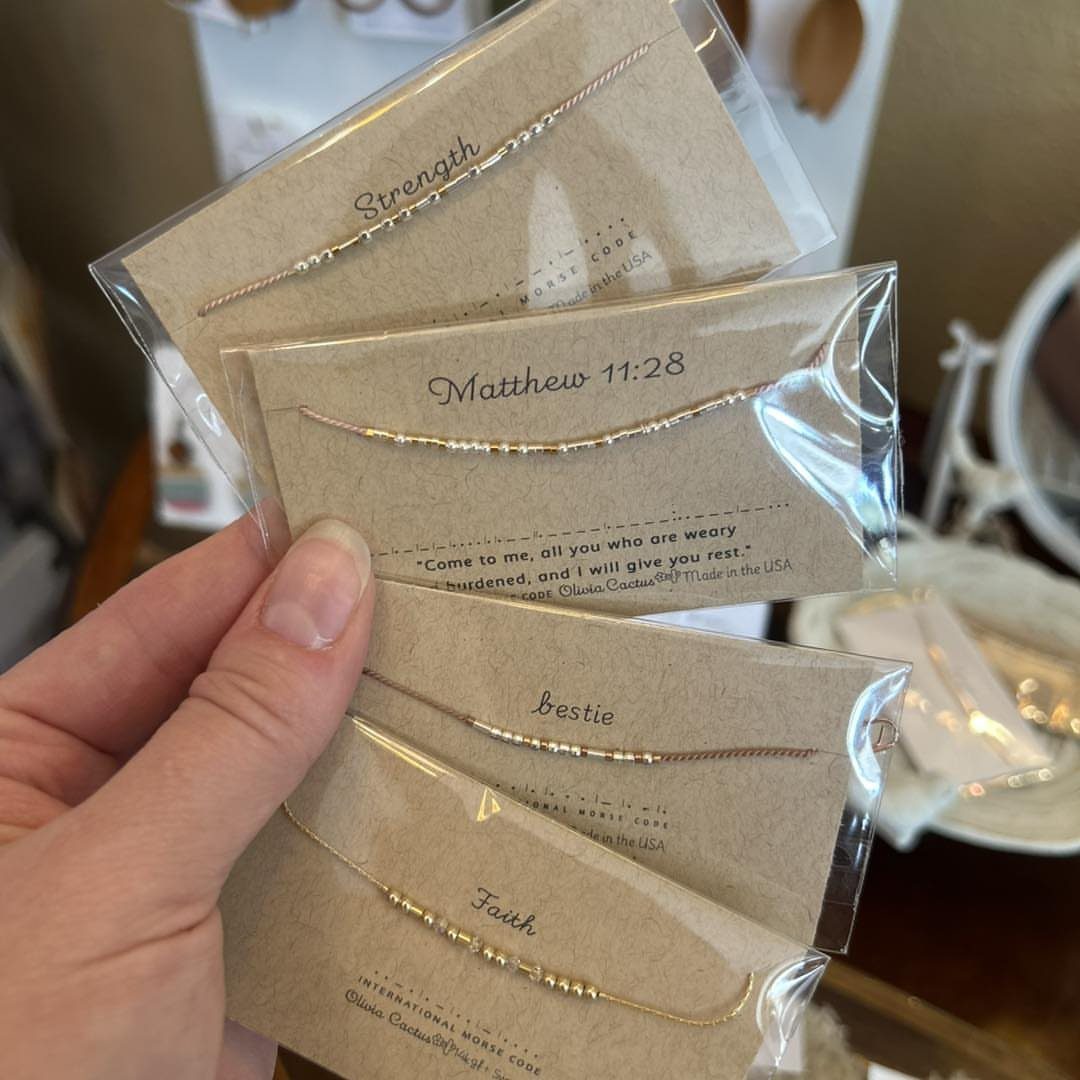 Gold Morse code bracelets on brown cards with words like Strength and Faith.