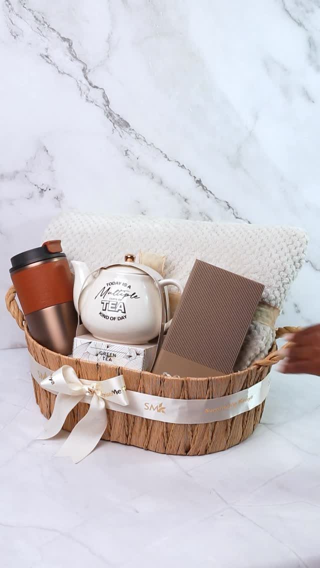 Woven gift basket with a teapot, travel mug, notebook, and tea on marble.