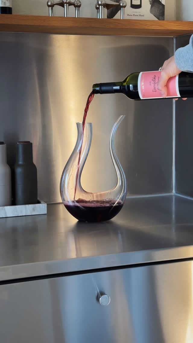 Red wine pouring from a bottle into a curved glass decanter on a counter