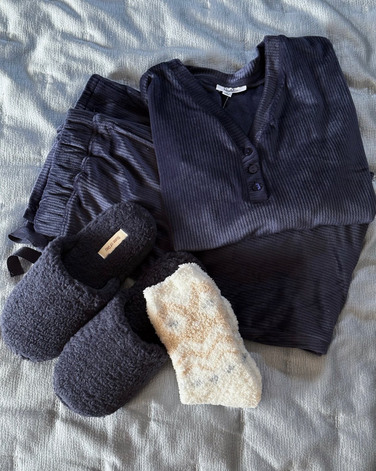 Navy blue ribbed pajama set with fuzzy slippers and a cream sock on grey fabric.