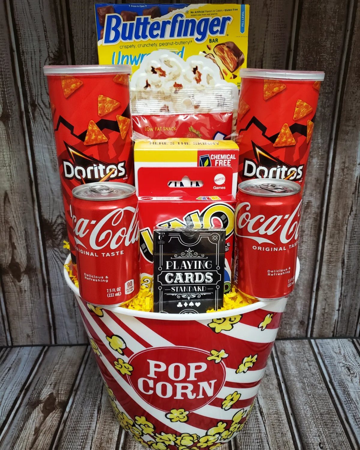 Popcorn bucket filled with Coca-Cola, Doritos, Butterfinger, and playing cards on wood.