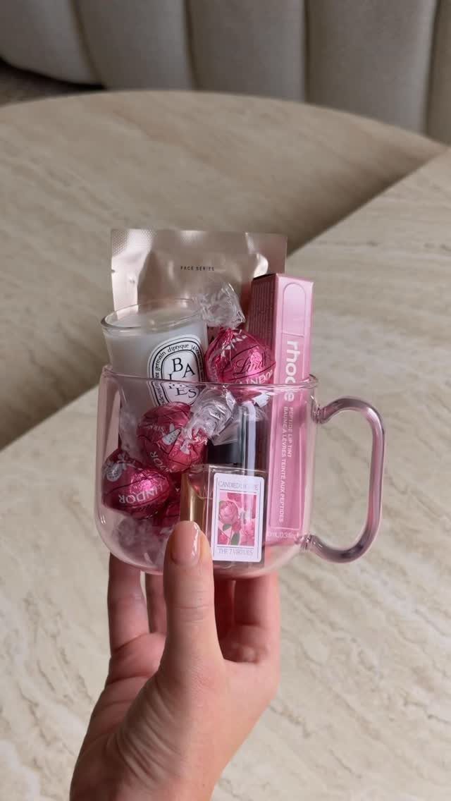 Clear glass mug filled with pink chocolates, a candle, and beauty products.