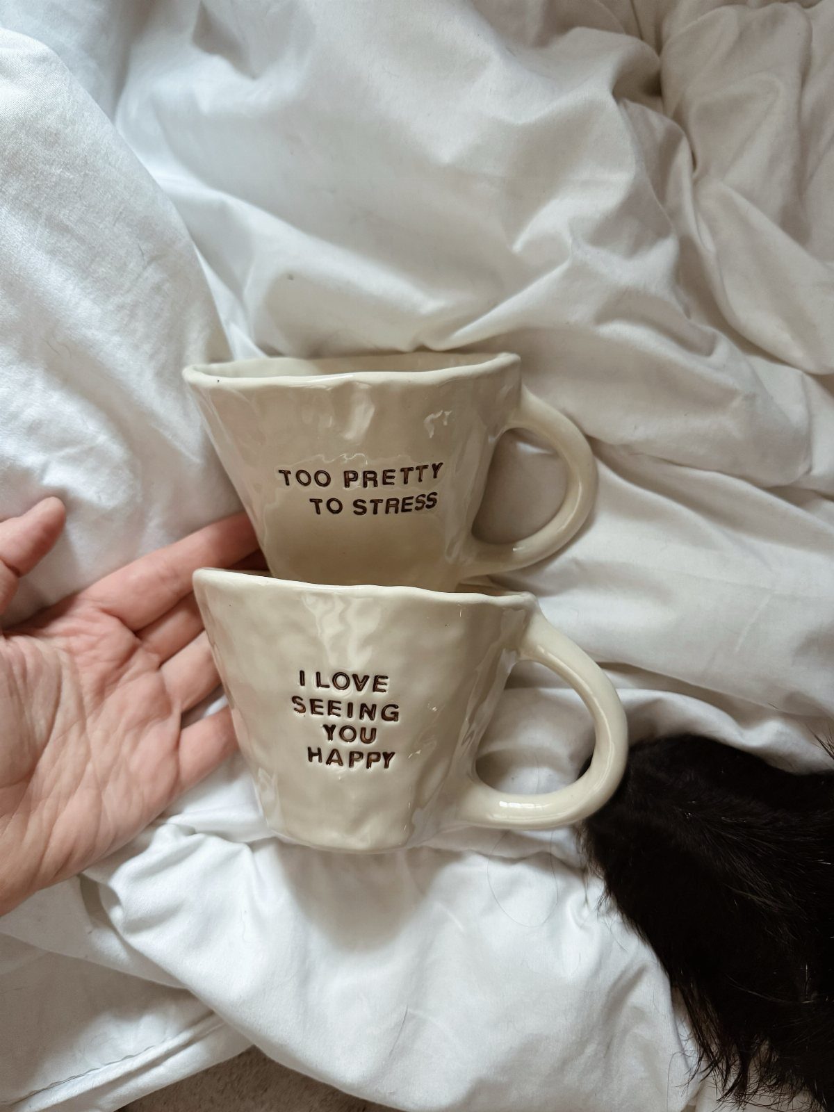 Two cream ceramic mugs with inspirational quotes resting on white wrinkled bedsheets.