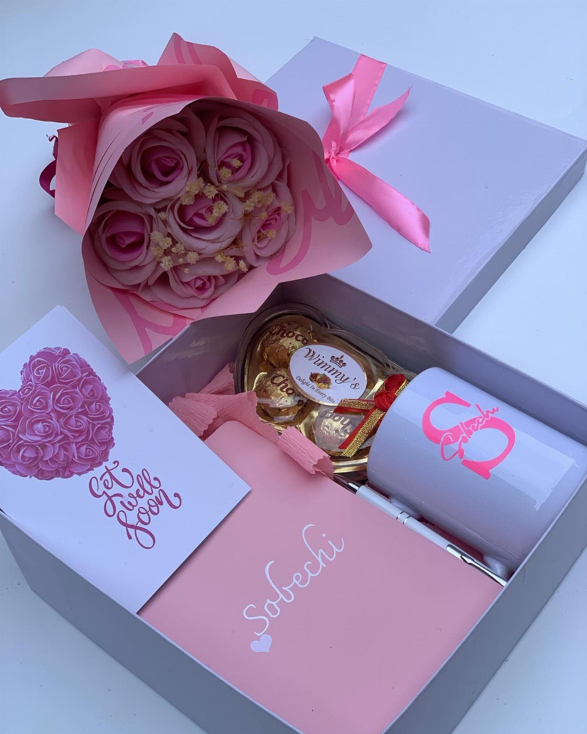 Gift box with a white mug, pink notebook, chocolates, and pink rose bouquet.