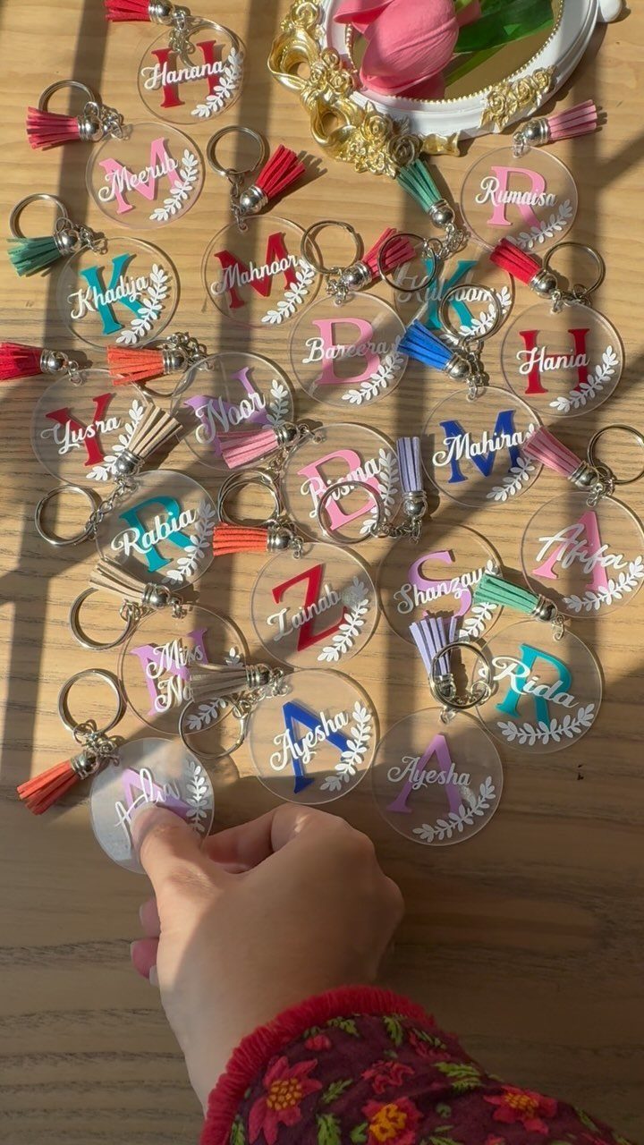 Circular acrylic keychains with colorful initials, names, and tassels on a wooden table