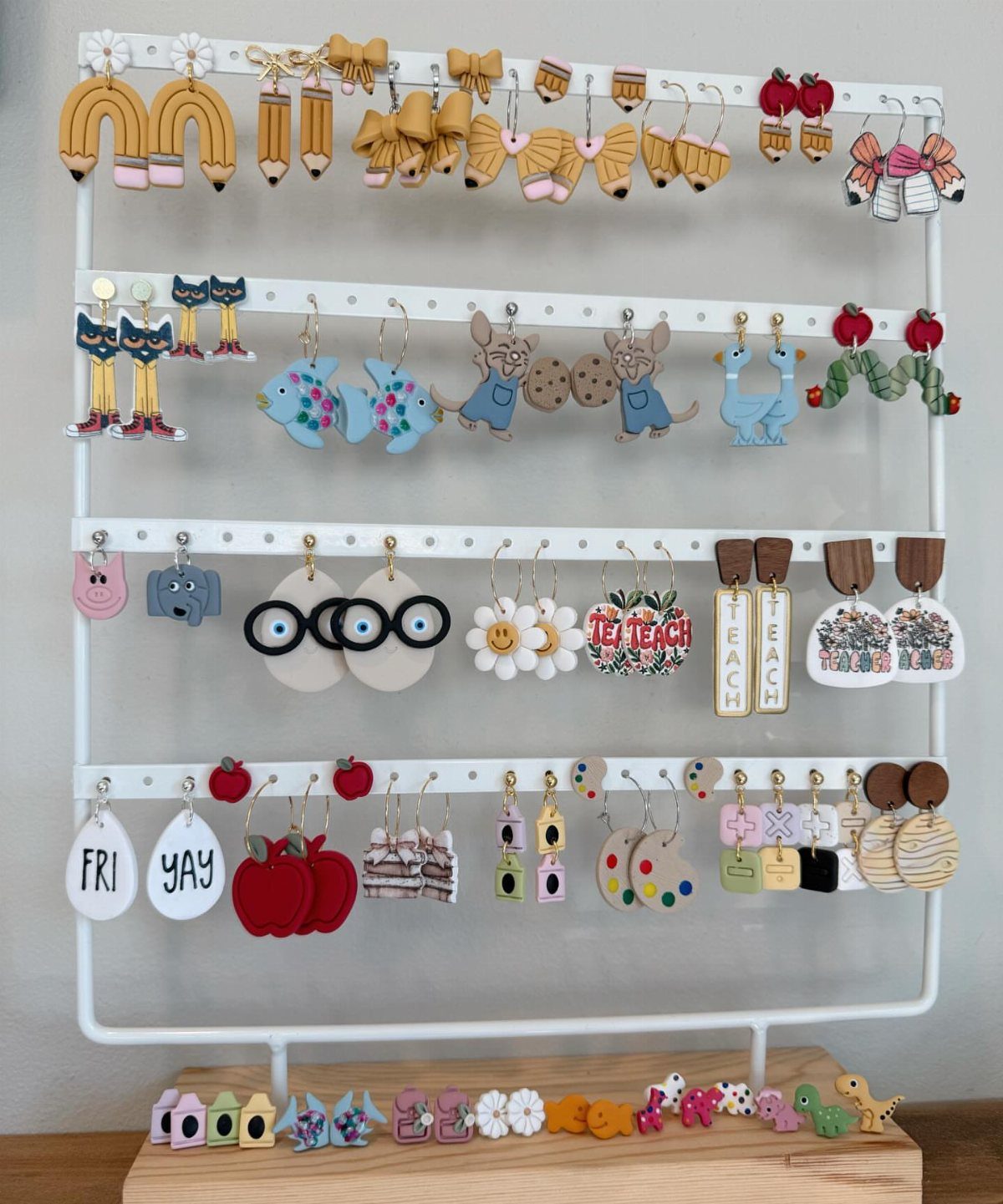 Teacher-themed earrings featuring pencils and apples displayed on a white metal rack.