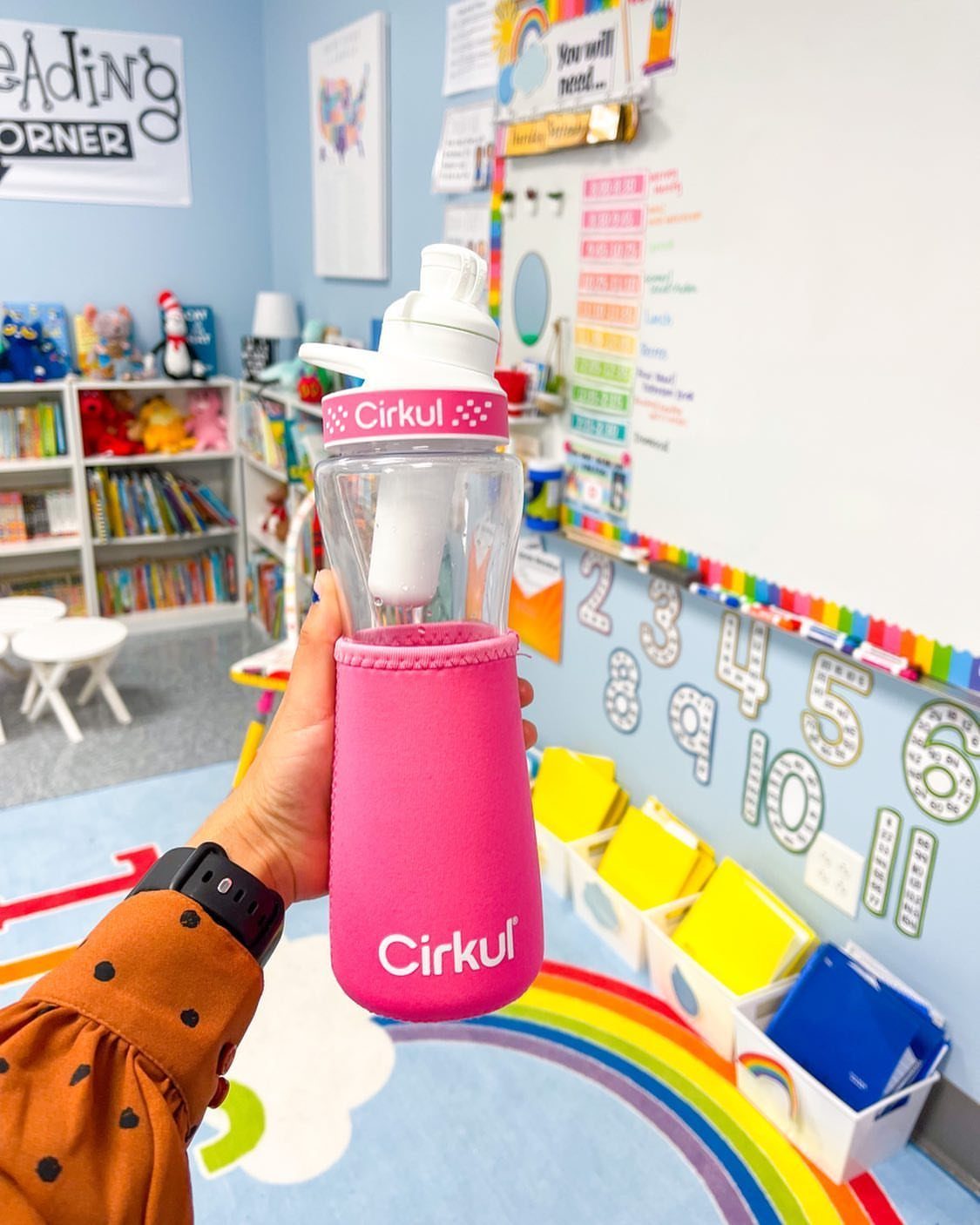 Cirkul water bottle with pink sleeve held in a colorful preschool classroom setting.