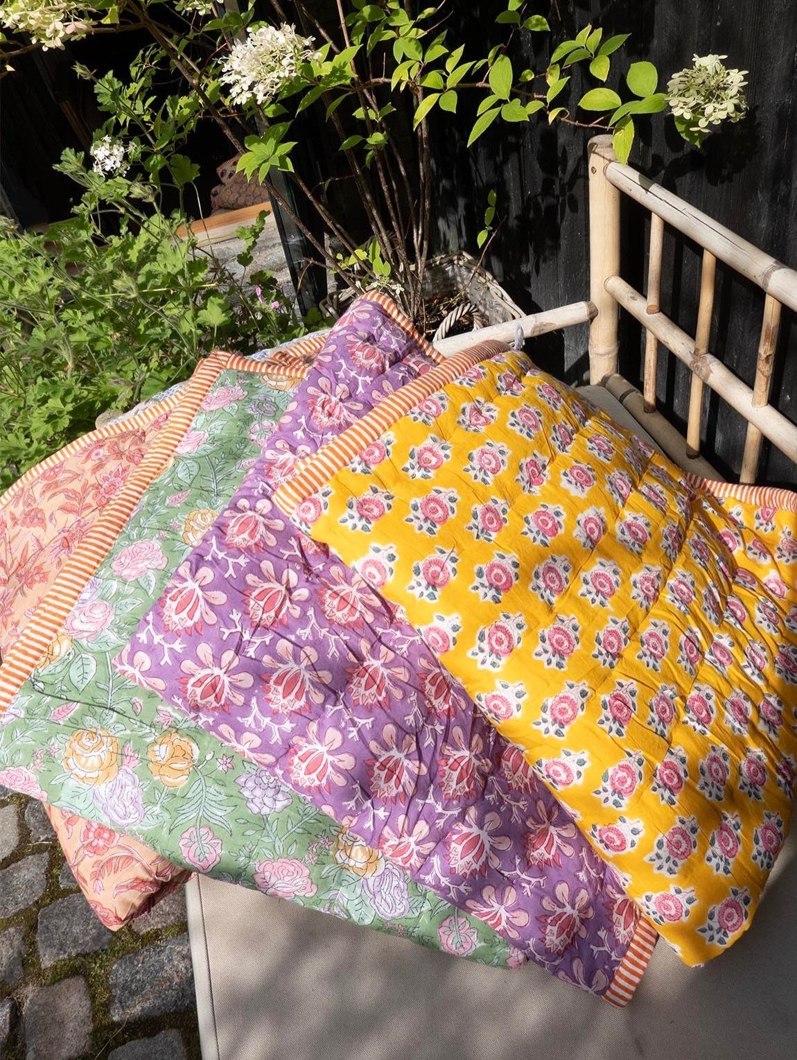 Colorful floral quilted blankets layered on an outdoor bench with garden greenery behind.