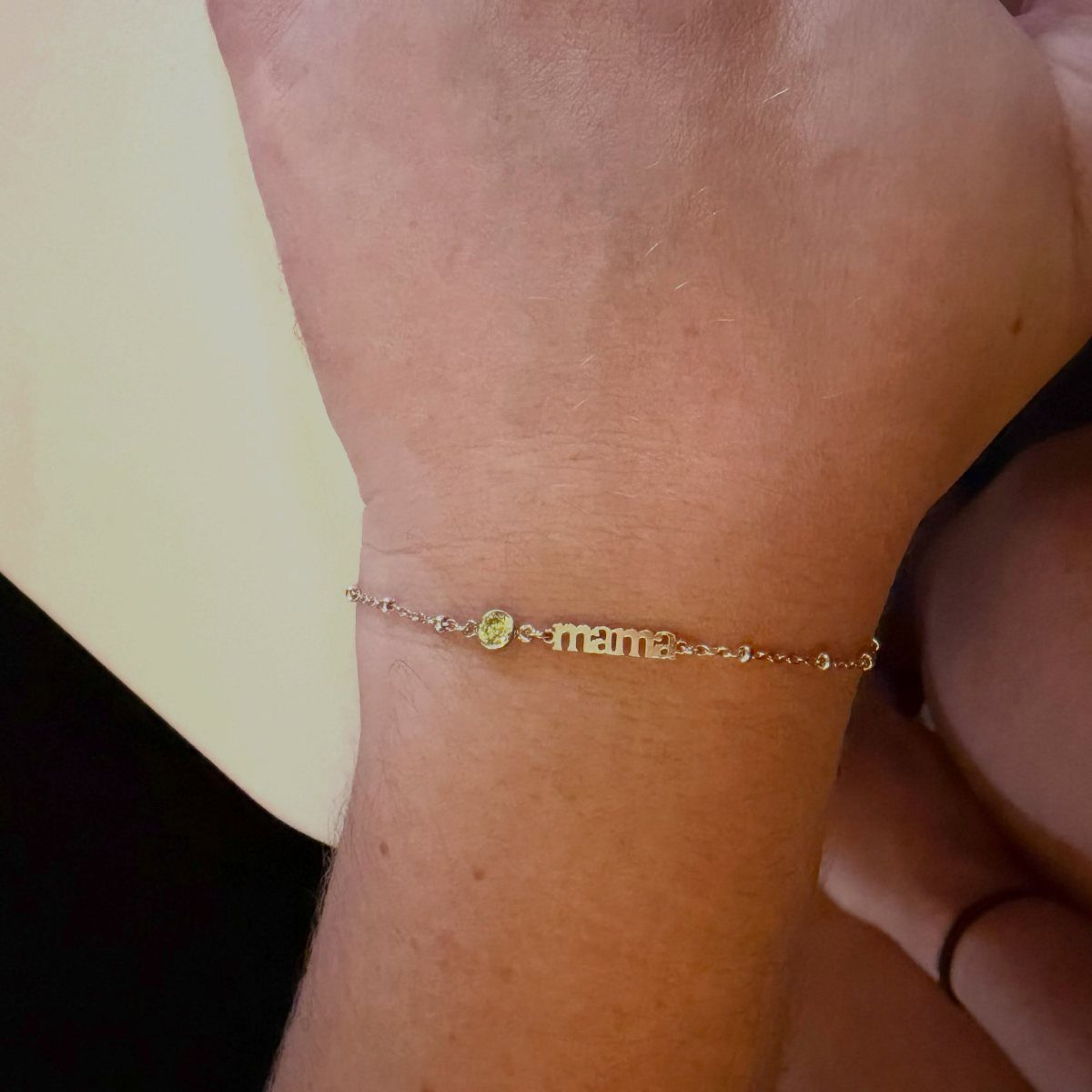 Gold chain bracelet with a green birthstone and 'mama' script on a wrist