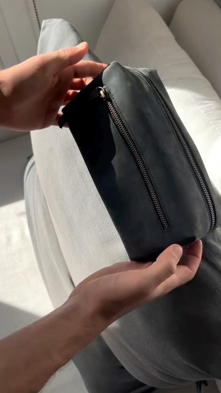 Black leather toiletry bag with zipper held by hands against grey trousers.