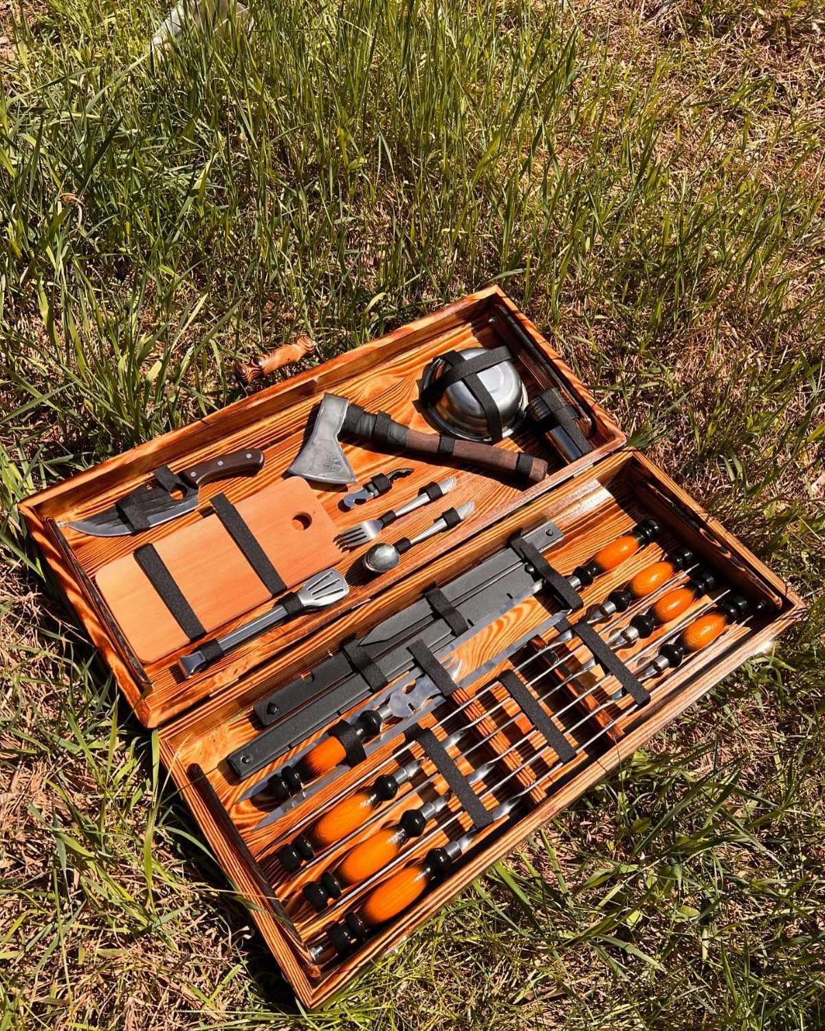 Wooden tool chest on grass containing an axe, knife, and cooking skewers.