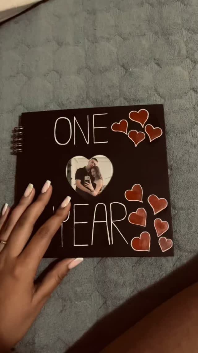 Hand holding a black anniversary scrapbook with a heart-shaped couple photo and red hearts.