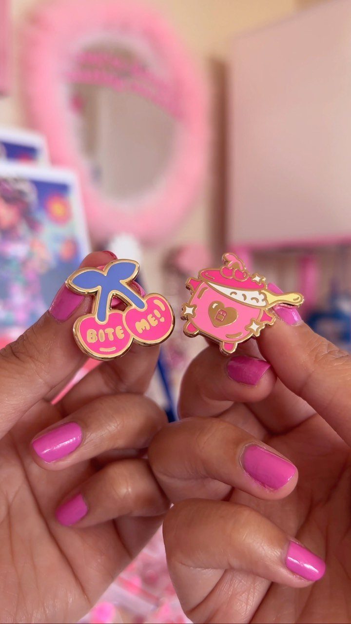 Two colorful enamel pins held by hands with bright pink nail polish.