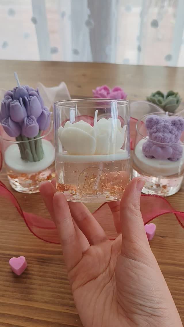 Hand holding a white flower-shaped candle in a clear glass holder on wood.