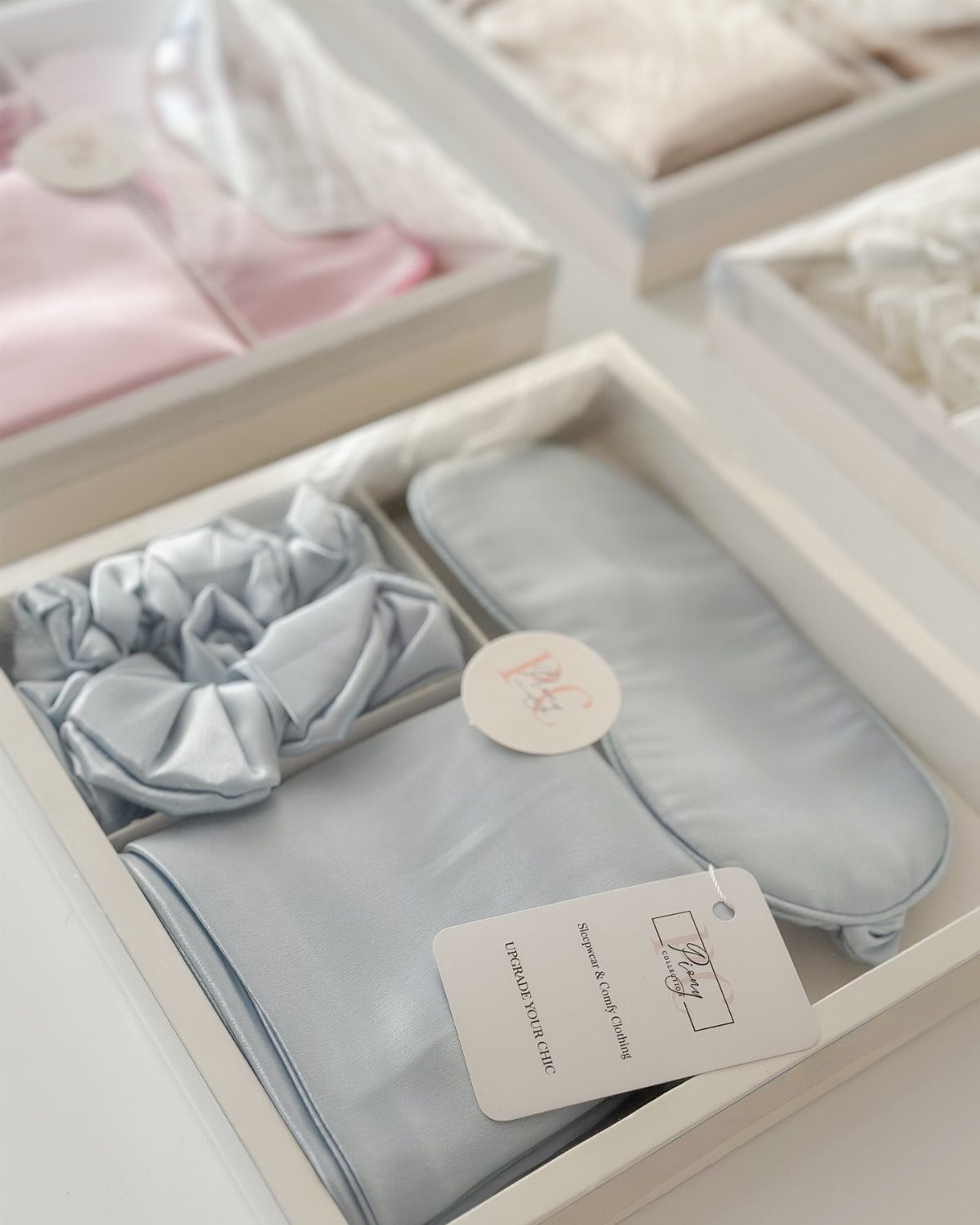 Light blue satin sleep mask, scrunchie, and pillowcase arranged in a white gift box.