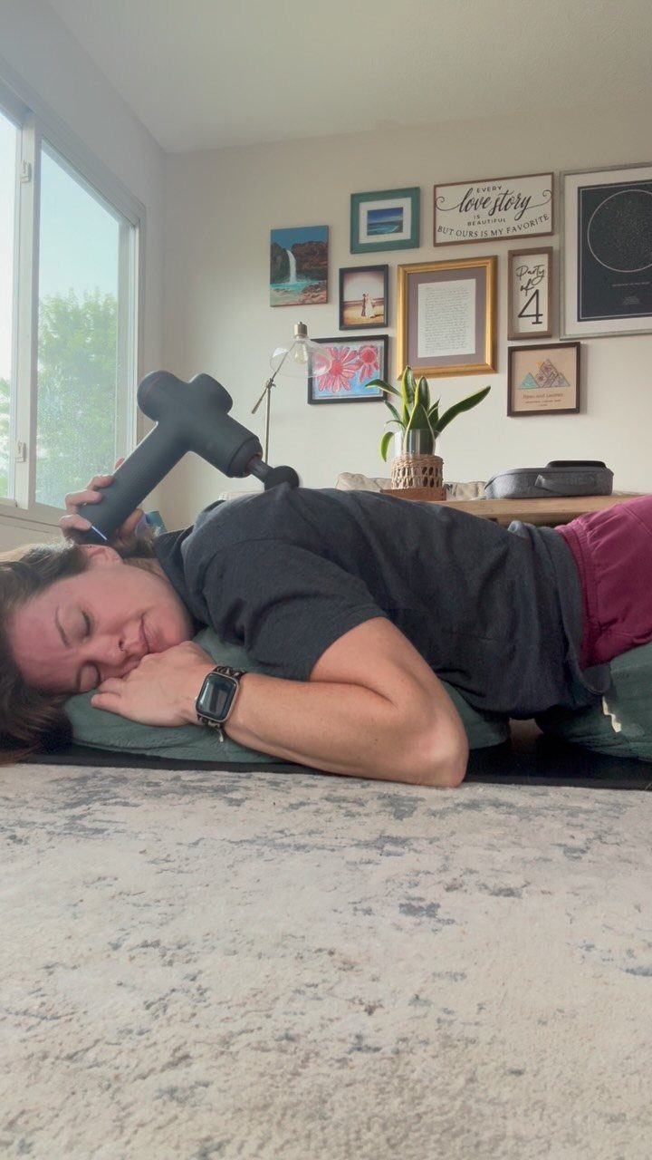 Woman using a black handheld massage gun on her shoulder while lying down.