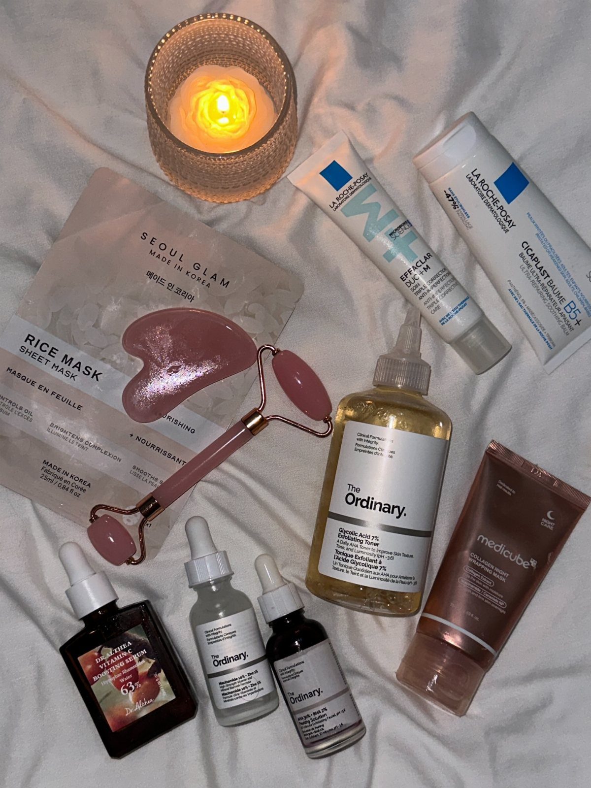 Skincare products, a facial roller, and a lit candle on white fabric.