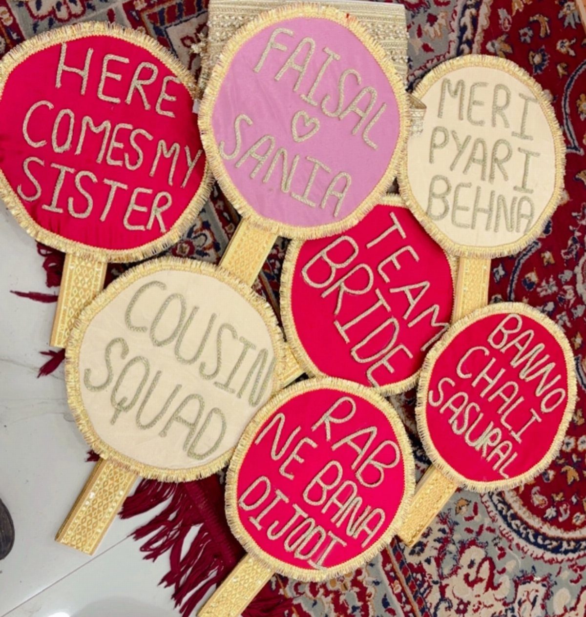 Embroidered handheld wedding fans with celebratory phrases resting on a red patterned rug.