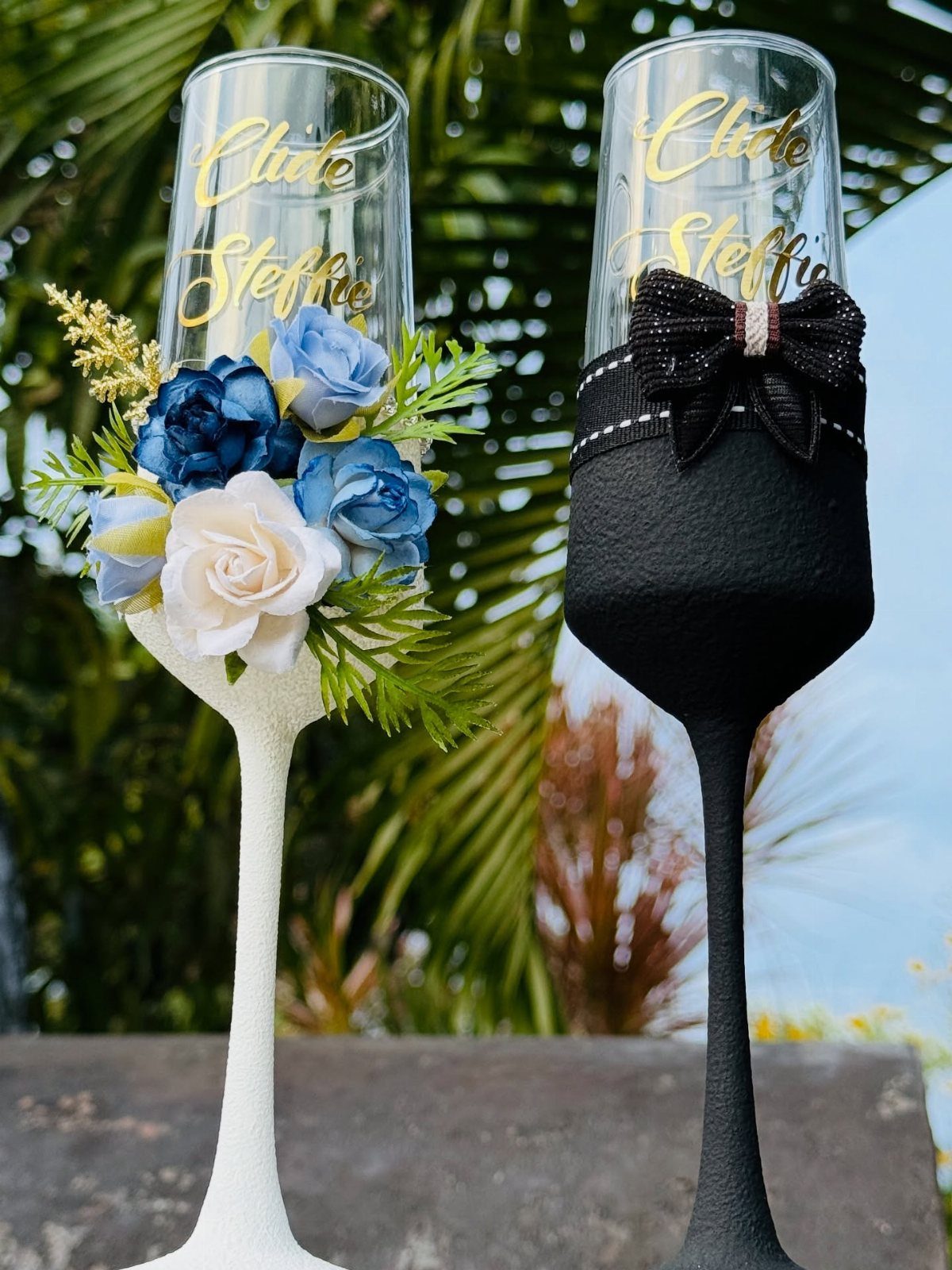 Two decorated champagne flutes, one with white roses and one with a black bow.