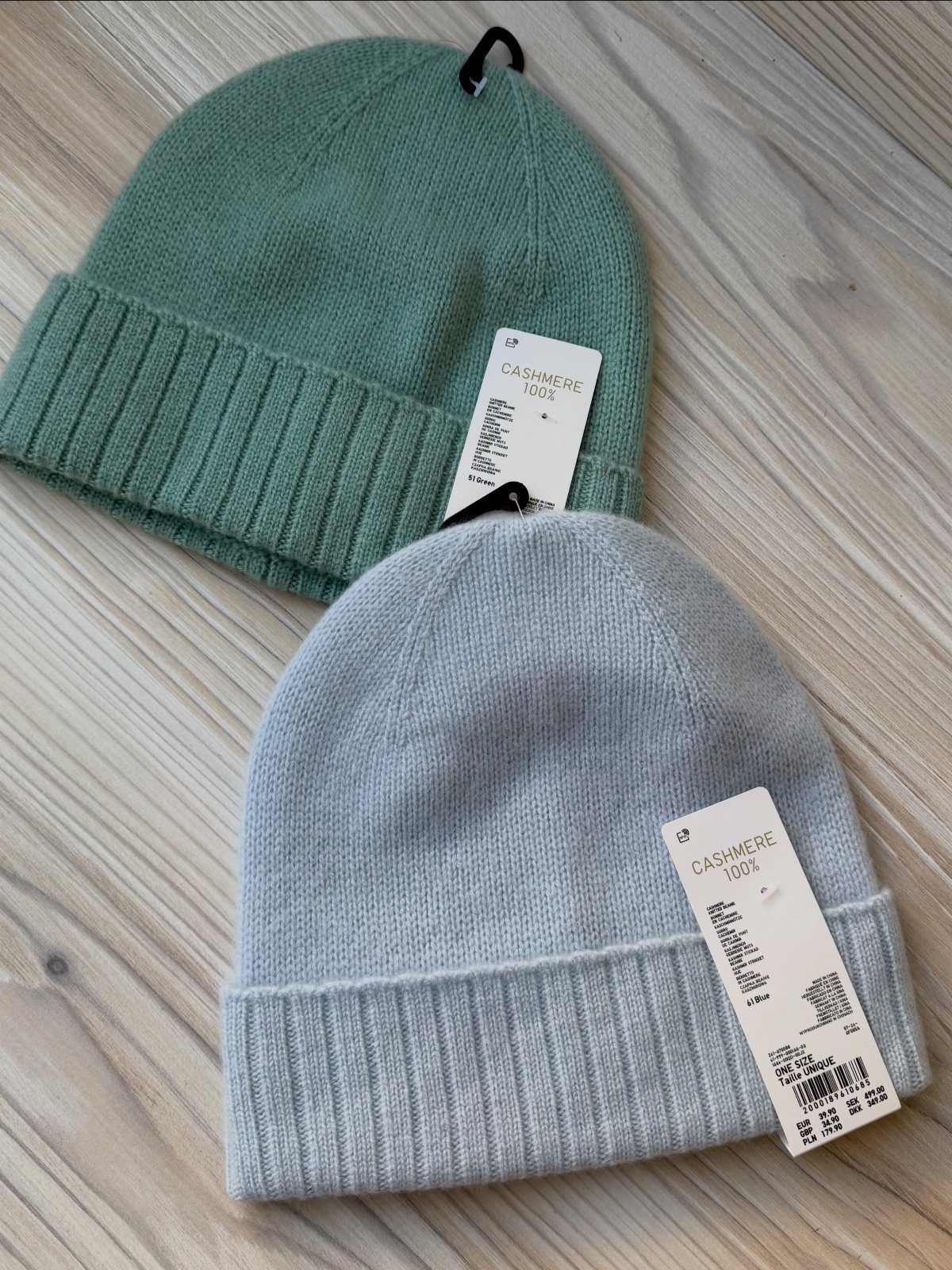 Two cashmere beanies in sage green and light blue on a wooden floor.