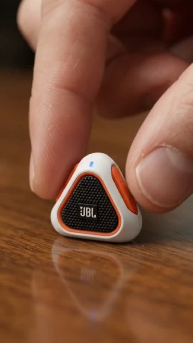 Small triangular JBL wireless speaker held by fingers on a wooden table