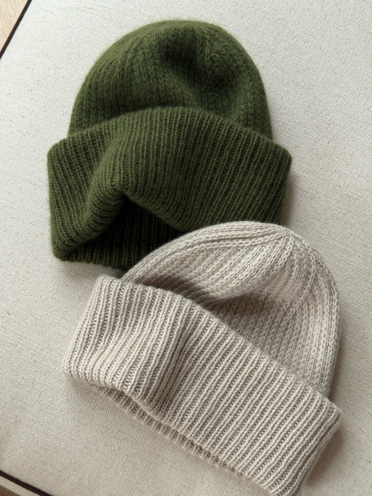 Olive green and light grey ribbed knit beanies on a neutral fabric background.