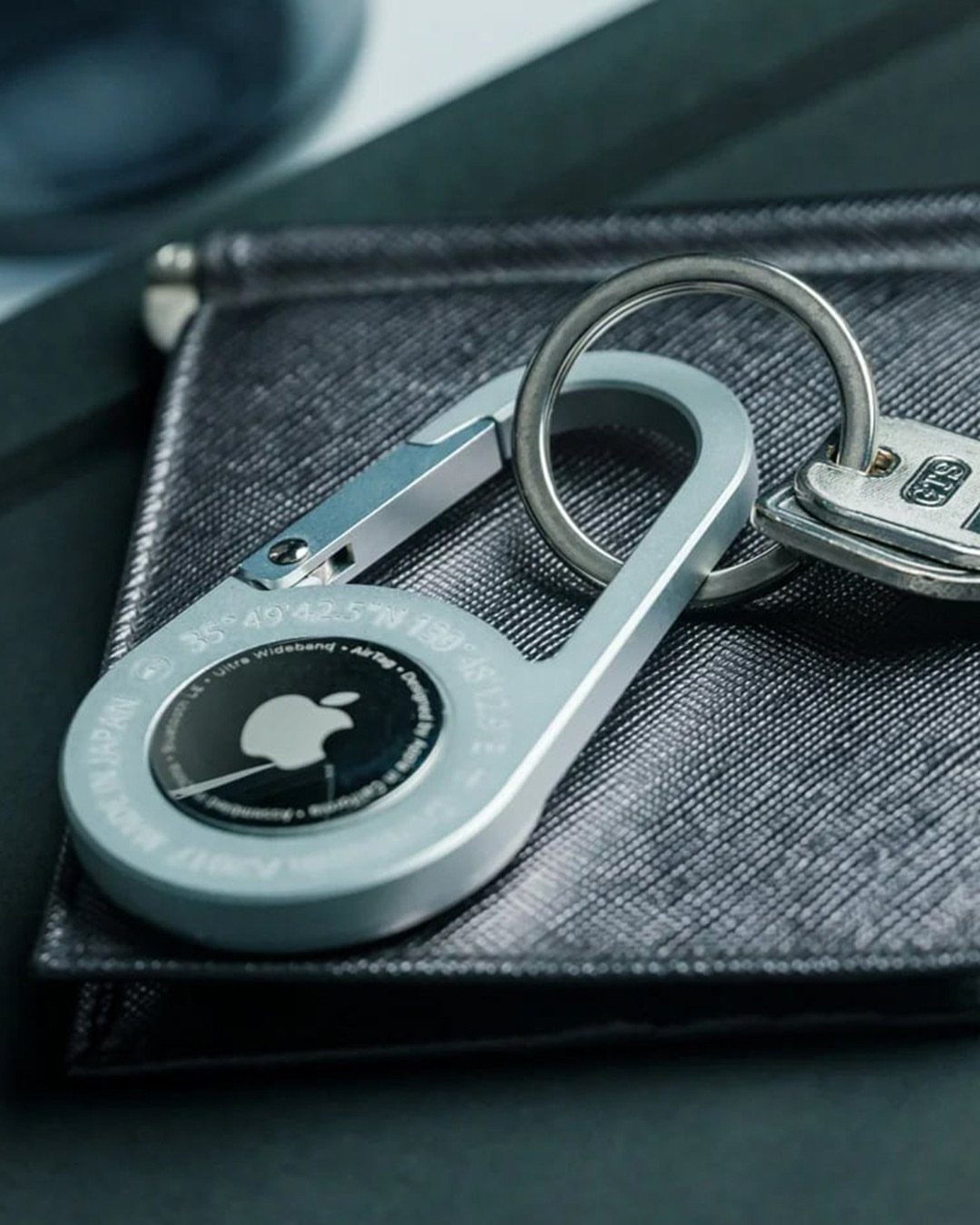 Apple AirTag in a silver keychain holder resting on a black leather wallet