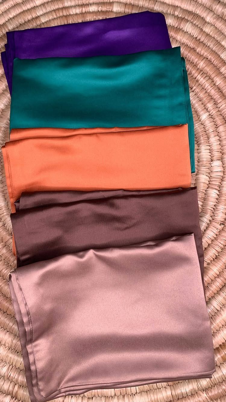 Five colorful satin pillowcases folded and stacked on a woven straw mat.