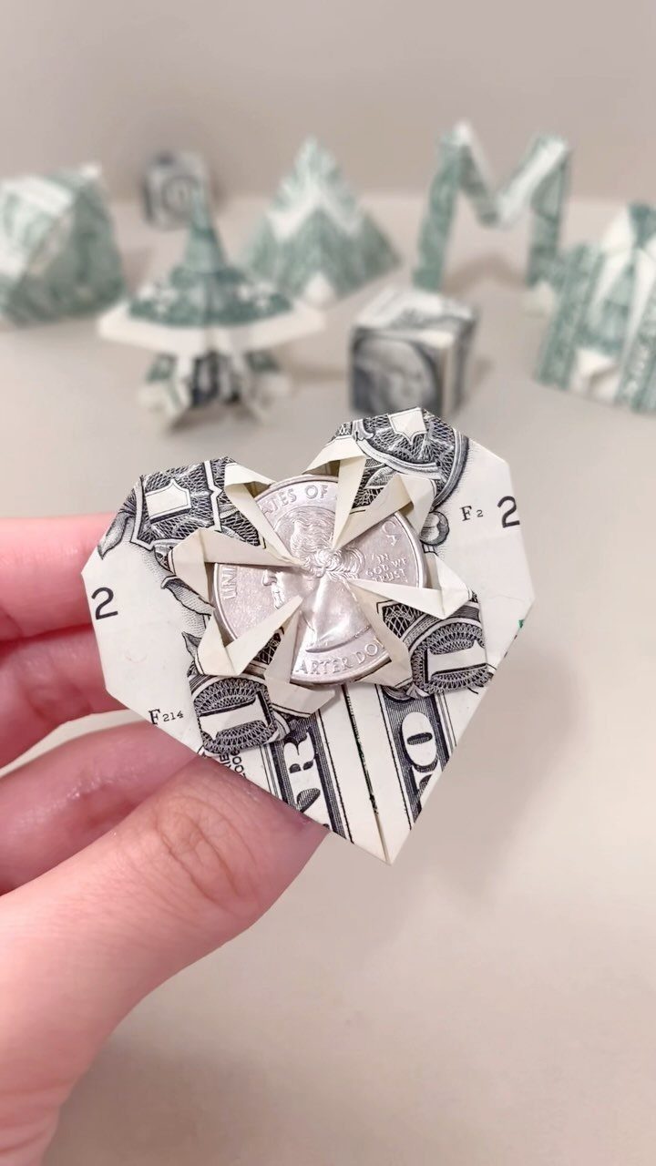 Heart-shaped origami folded from dollar bills held by a hand with a coin center