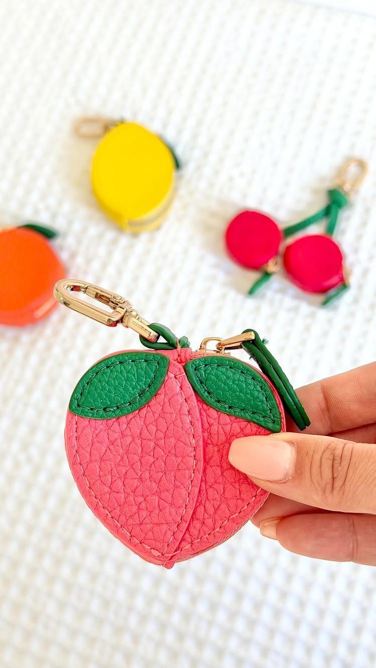 Pink peach-shaped coin purse held by a hand with other fruit purses nearby.