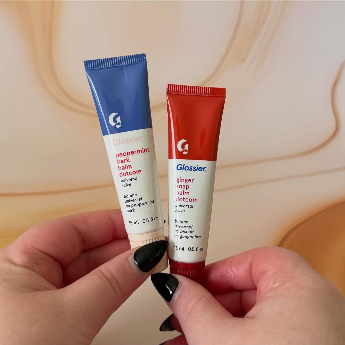 Two Glossier lip balm tubes held by hands with black nail polish.