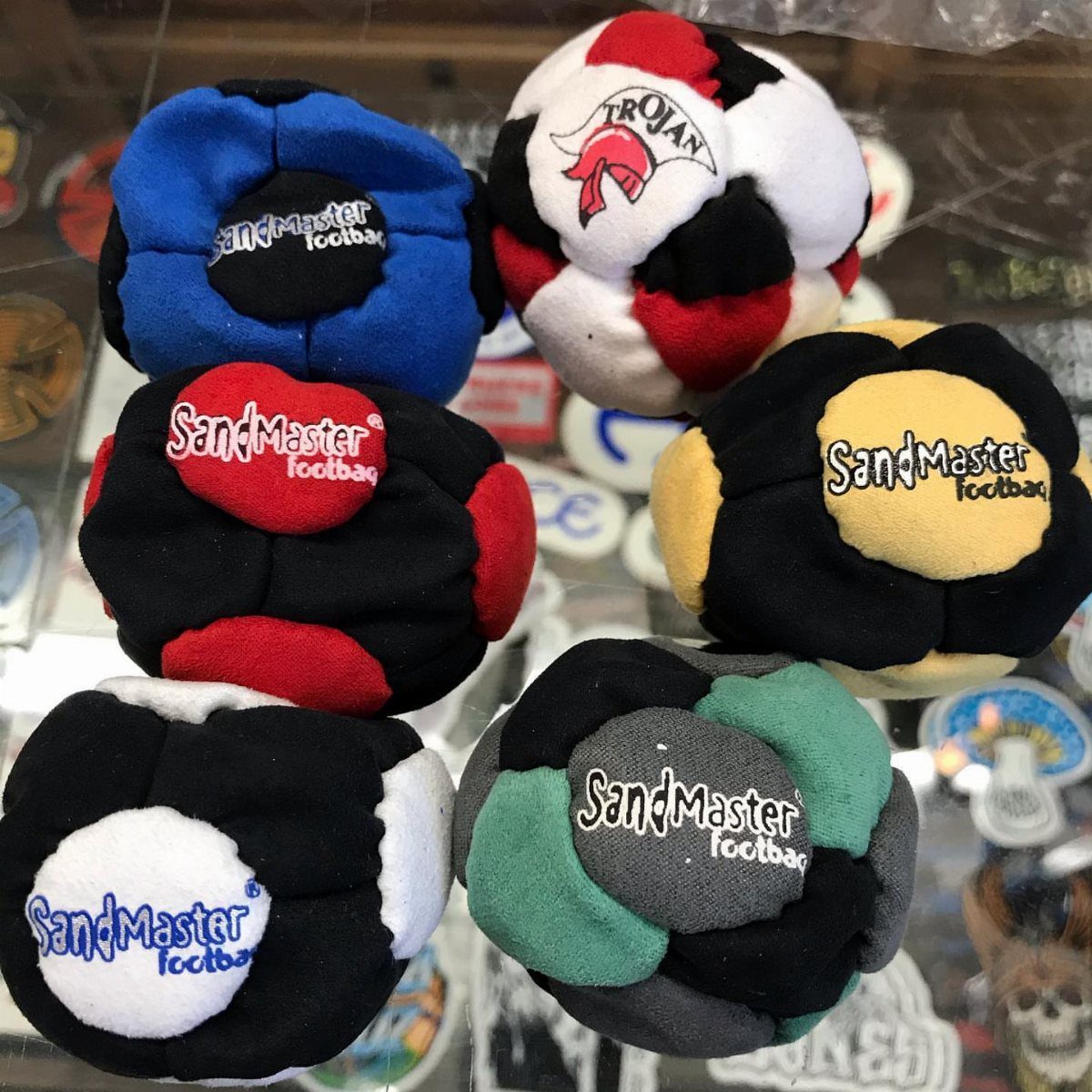 Colorful SandMaster footbags piled together on a glass display surface.