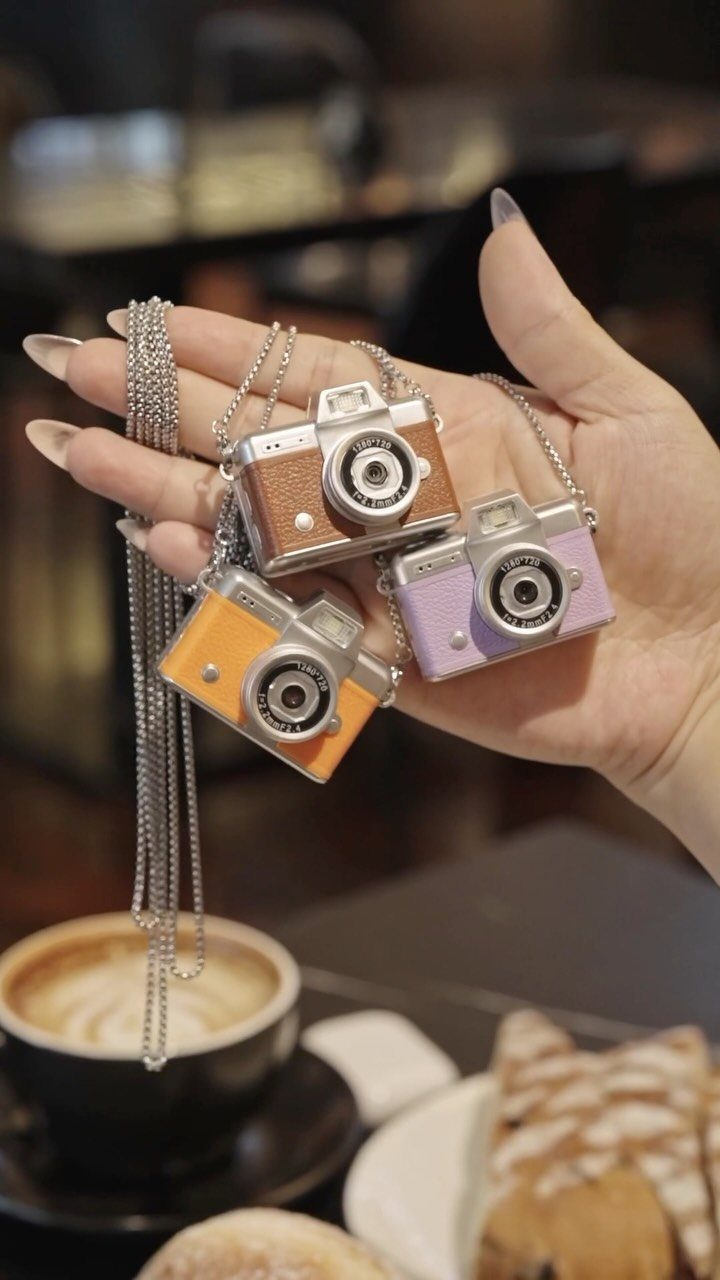 Three small camera pendants in brown, orange, and purple held in a hand.