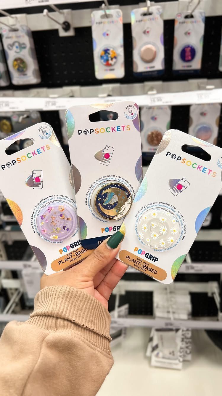 Hand holding three packaged PopSockets phone grips in a retail store setting.
