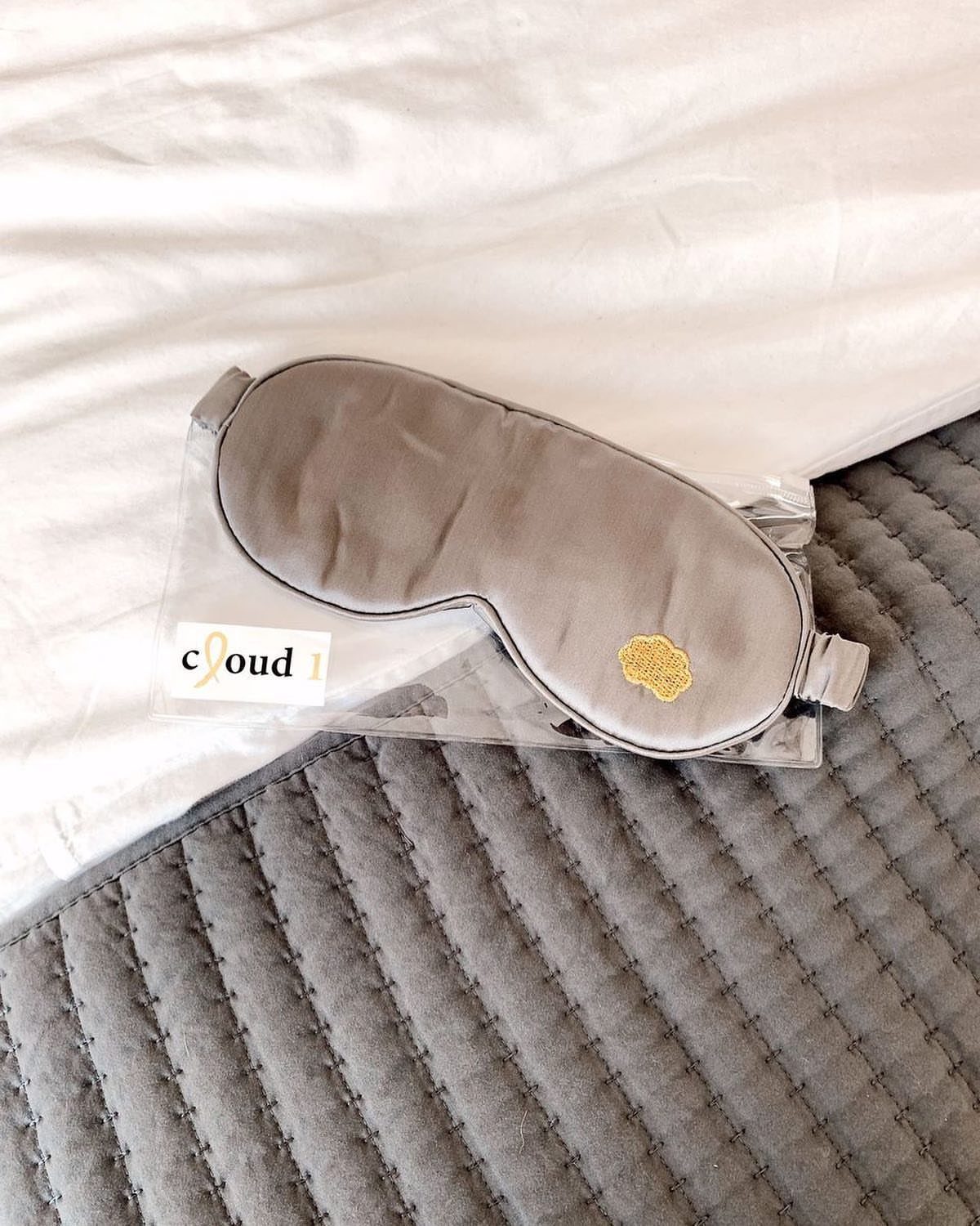 Grey satin sleep mask in clear packaging on white and grey bedding.