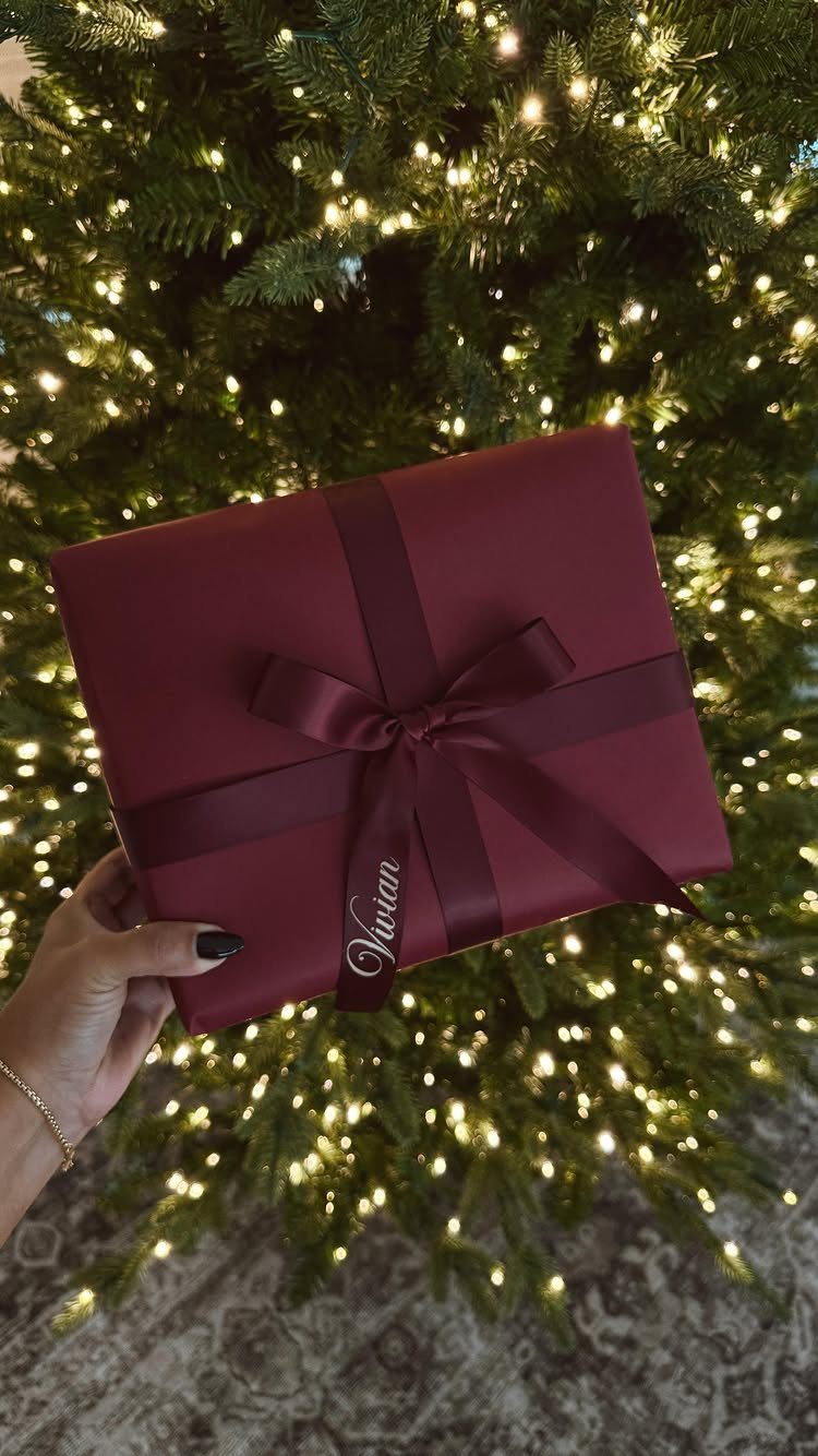 Burgundy wrapped gift box with a monogrammed ribbon held before a lit Christmas tree