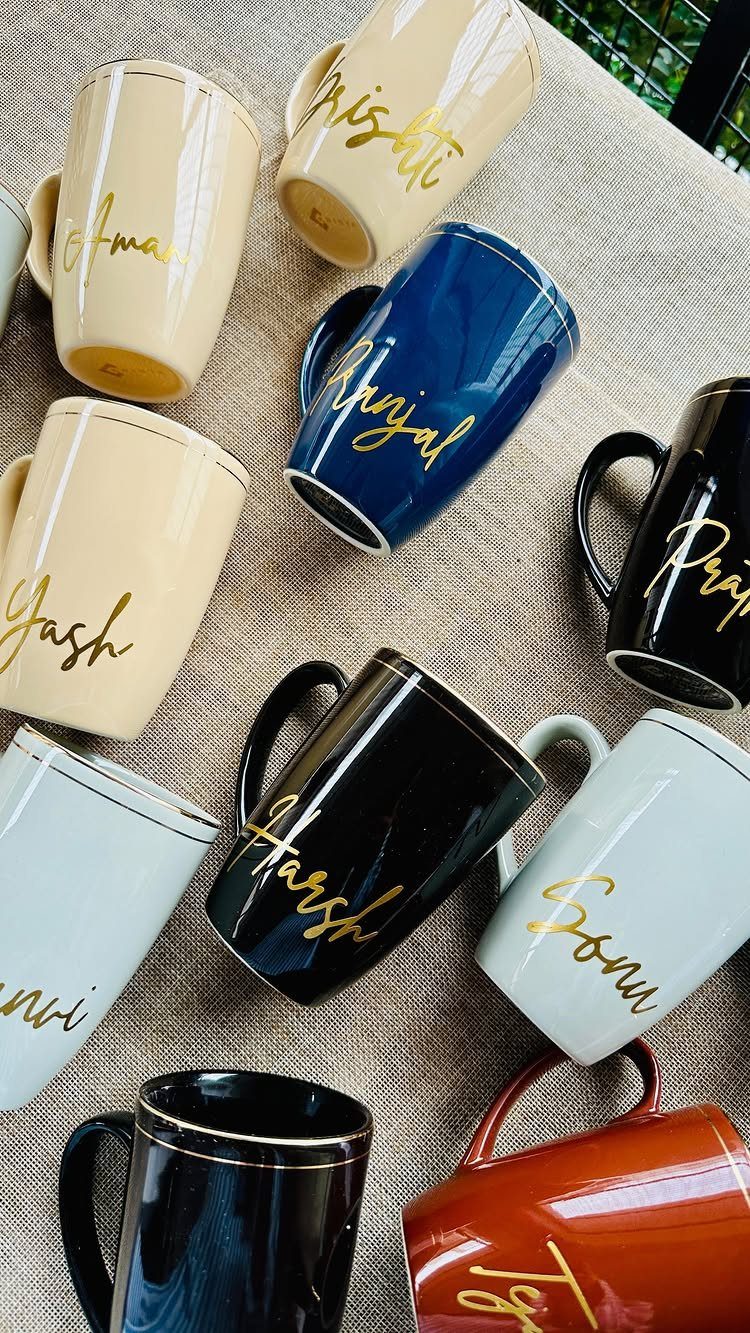 Assorted colorful ceramic coffee mugs with personalized names written in gold cursive script.