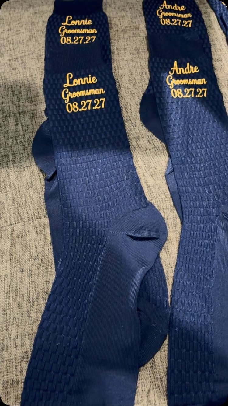 Navy blue groomsman socks with gold personalized names and dates on grey fabric.