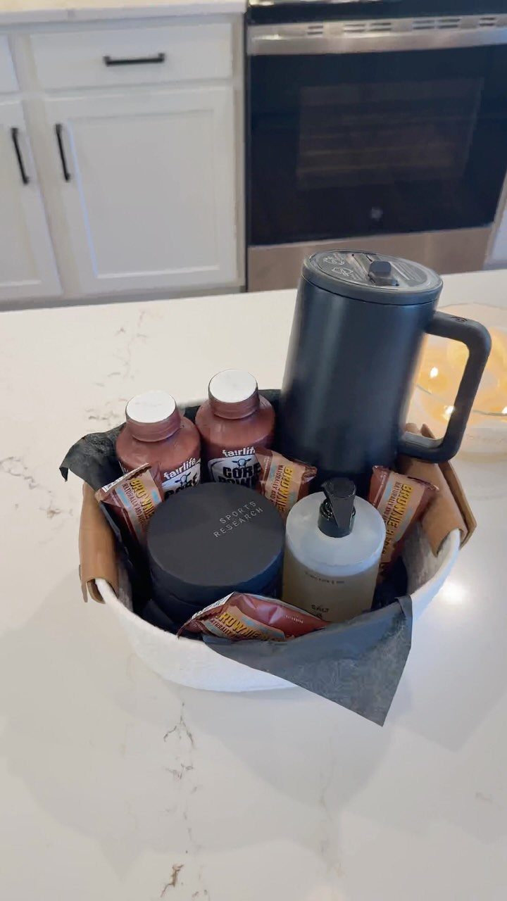 White gift basket with travel mug, snacks, and toiletries on a marble counter.