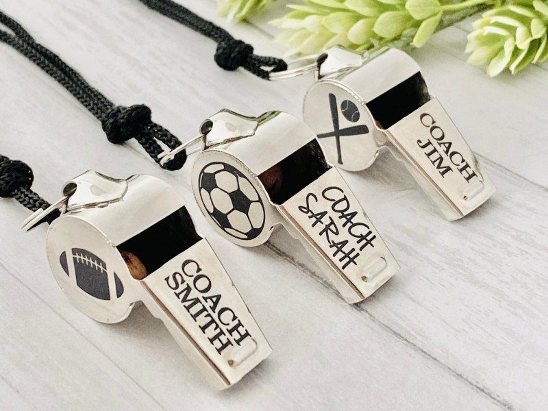 Three personalized silver metal whistles engraved with sports icons and coach names.
