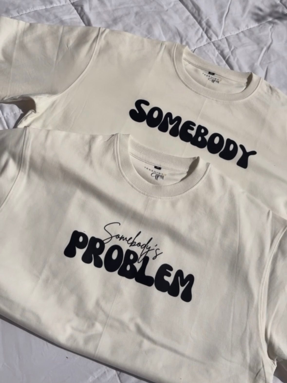 Two cream sweatshirts with black text reading Somebody and Somebody's Problem on white fabric